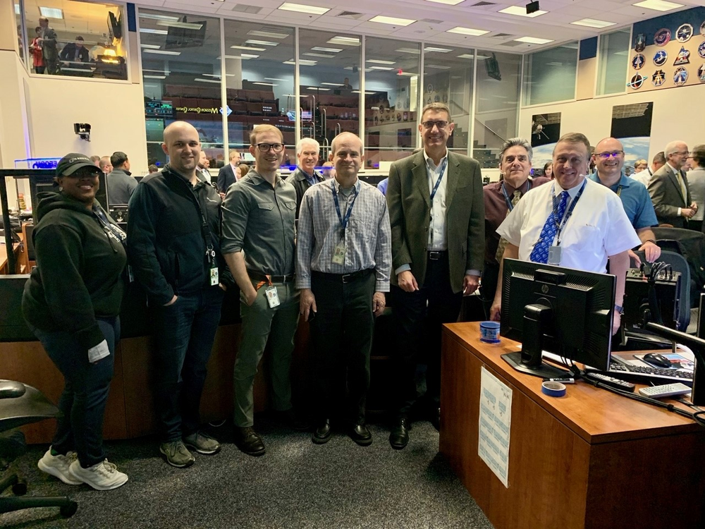 A group of men and women stand together in a flight control room.